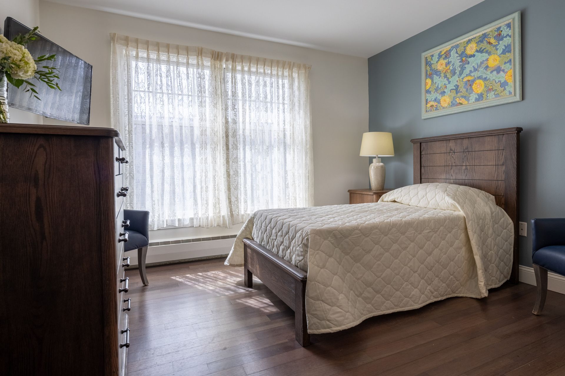Bedroom with bed, dresser, window, blue accent wall, art, lamp.
