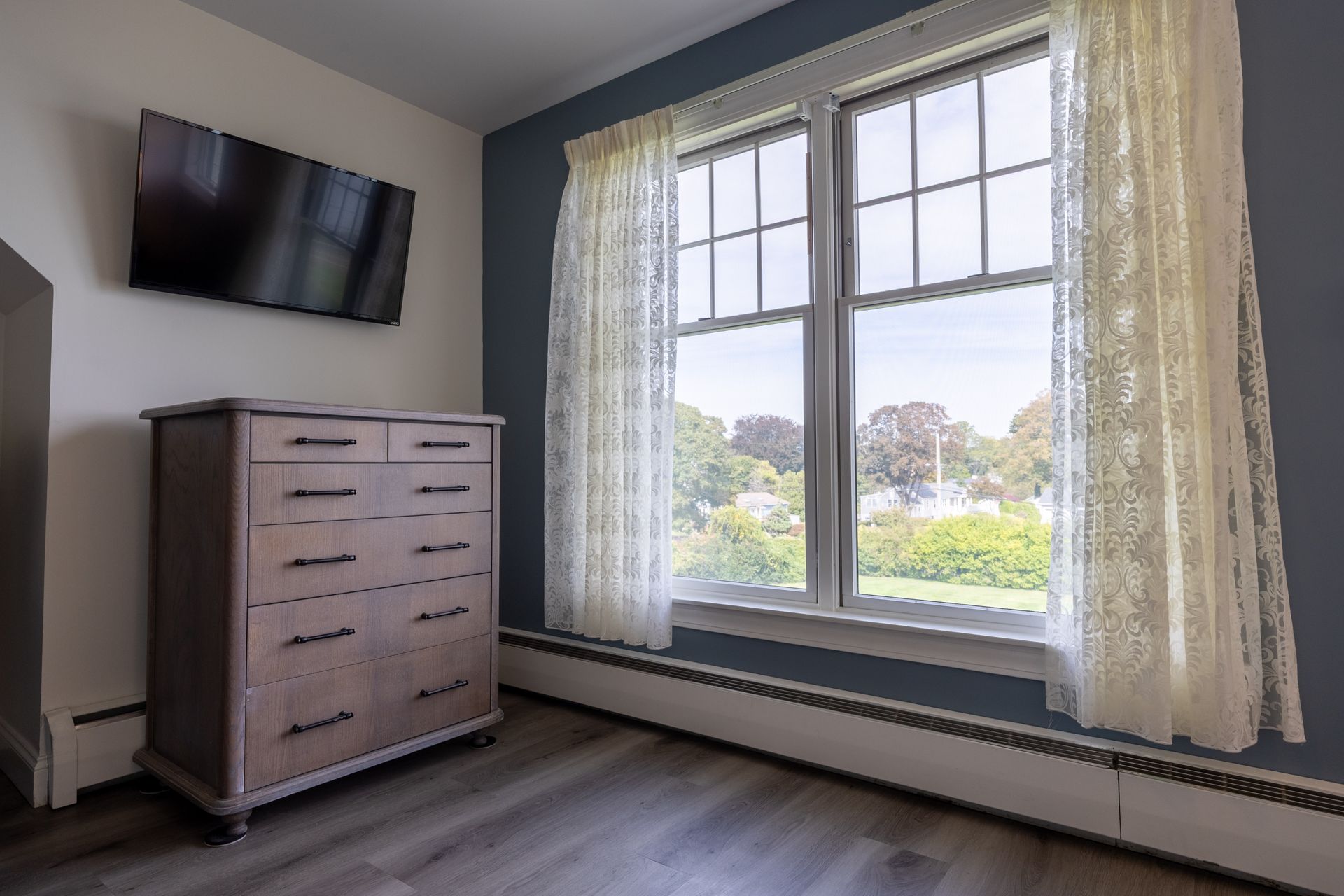 Bedroom with a dresser, window, TV, and lace curtains. Blue wall.