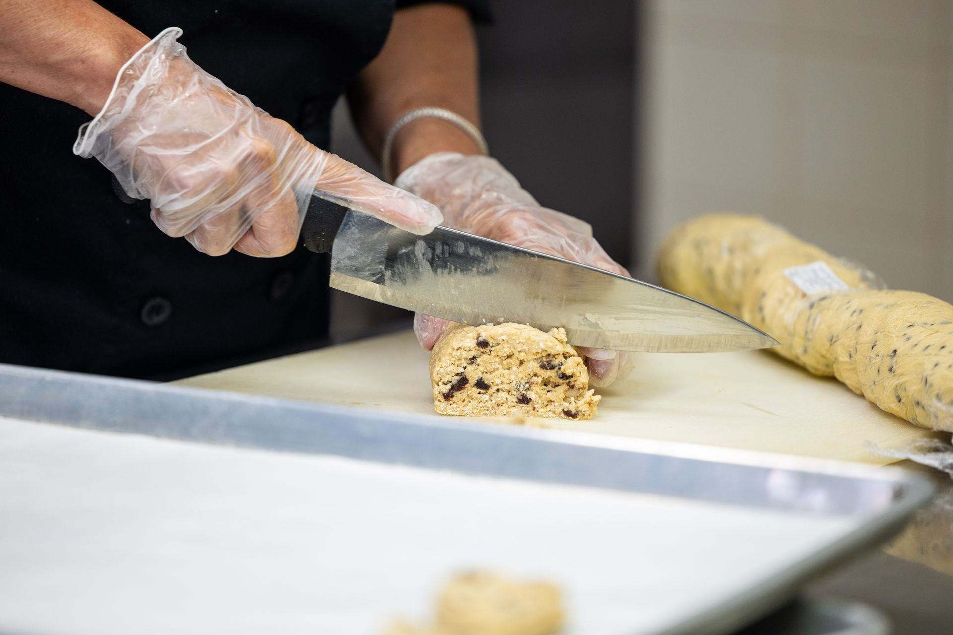 Person in gloves slicing cookie dough log on a cutting board, near a baking sheet.