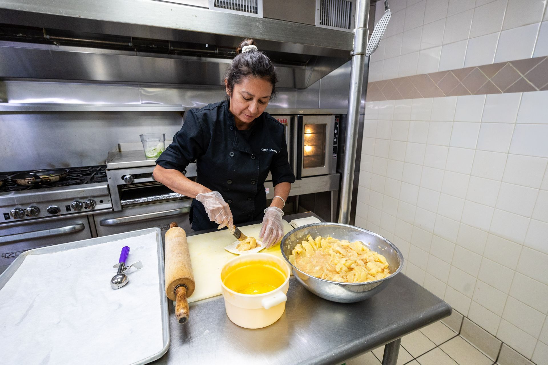 A chef in a black uniform cuts food at a stainless steel table in a commercial kitchen.