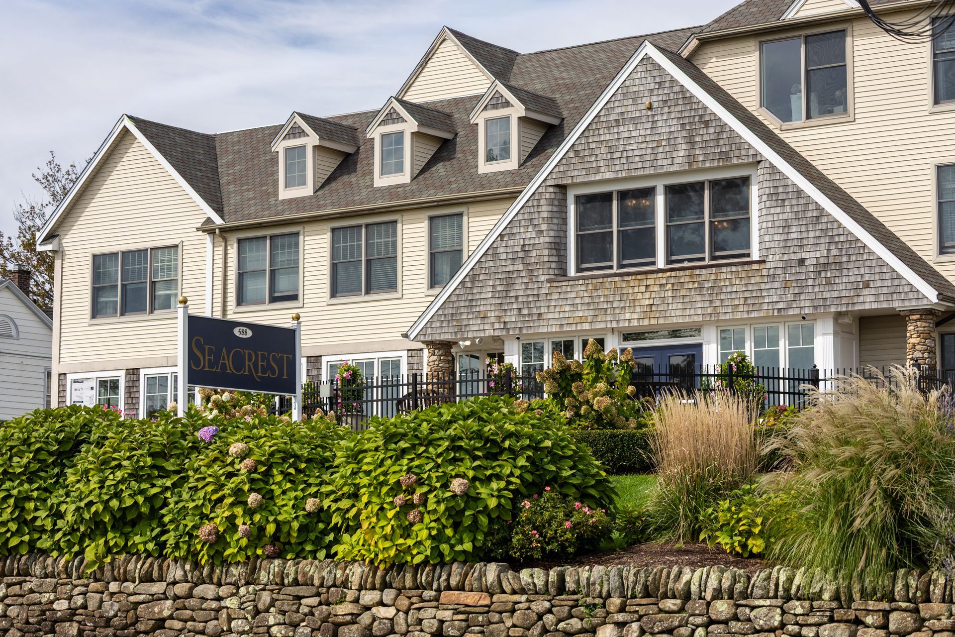 Seaglass Inn, light-colored building with multiple windows, stone wall and lush bushes in front, overcast sky.