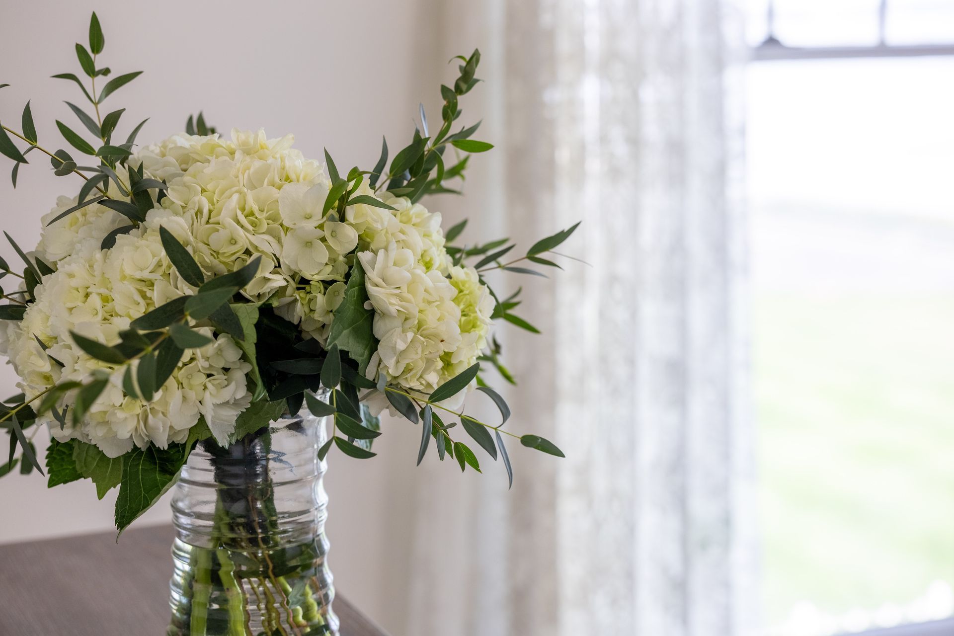 White hydrangea bouquet in a glass vase, greenery accents, beside a lace curtained window.