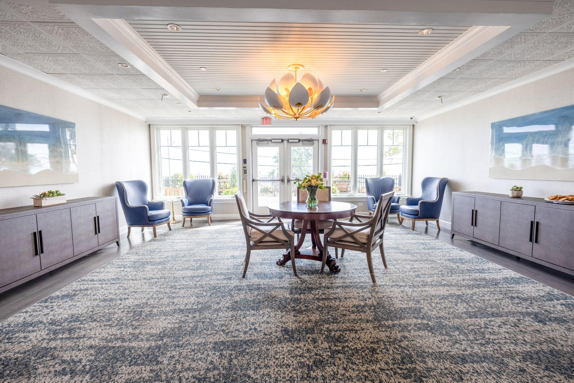 Bright room with round table, chairs, blue armchairs, and cabinets. Large rug and floral chandelier.