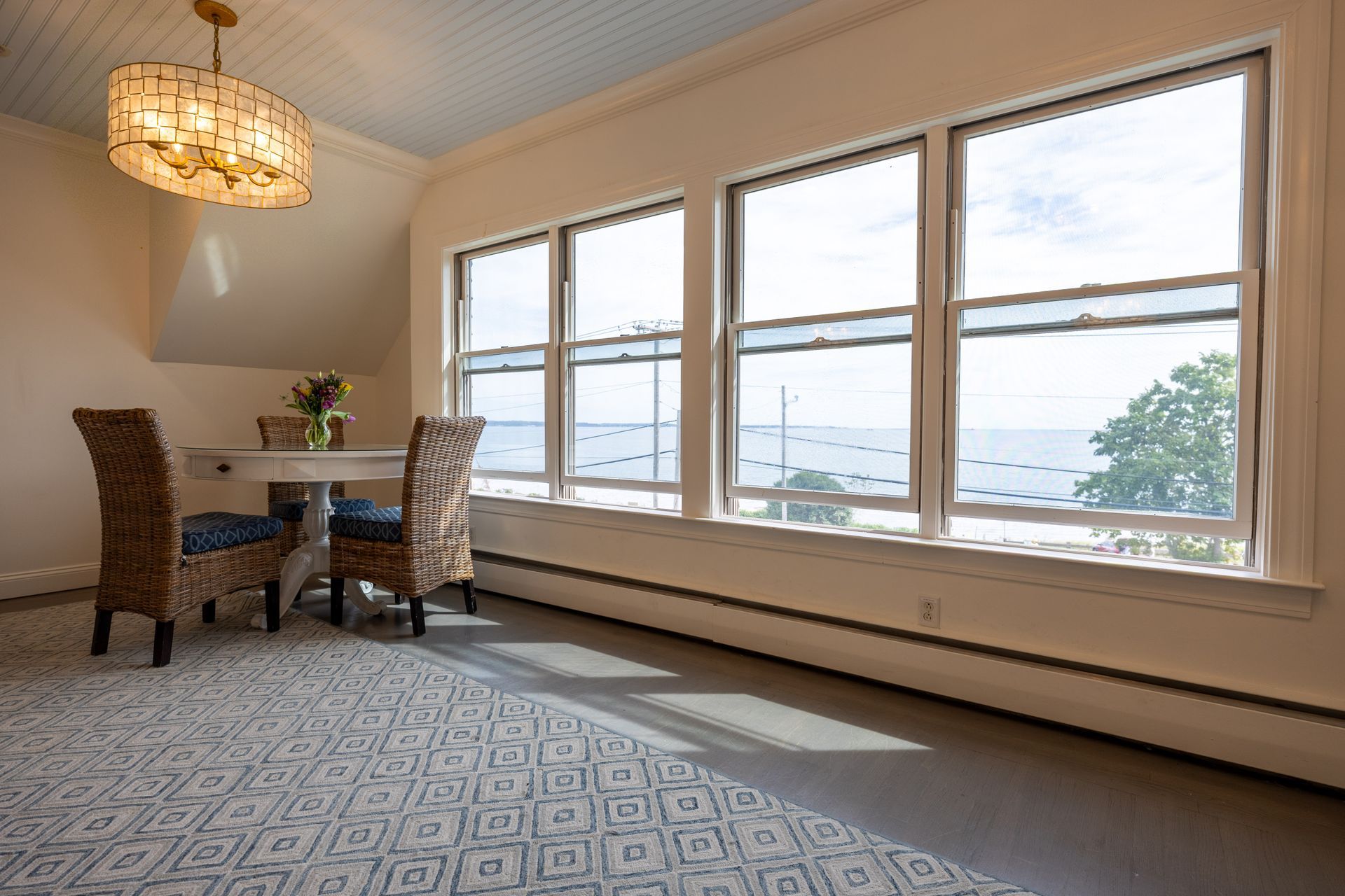 Dining area with water view: white table, wicker chairs, large windows, and patterned rug.