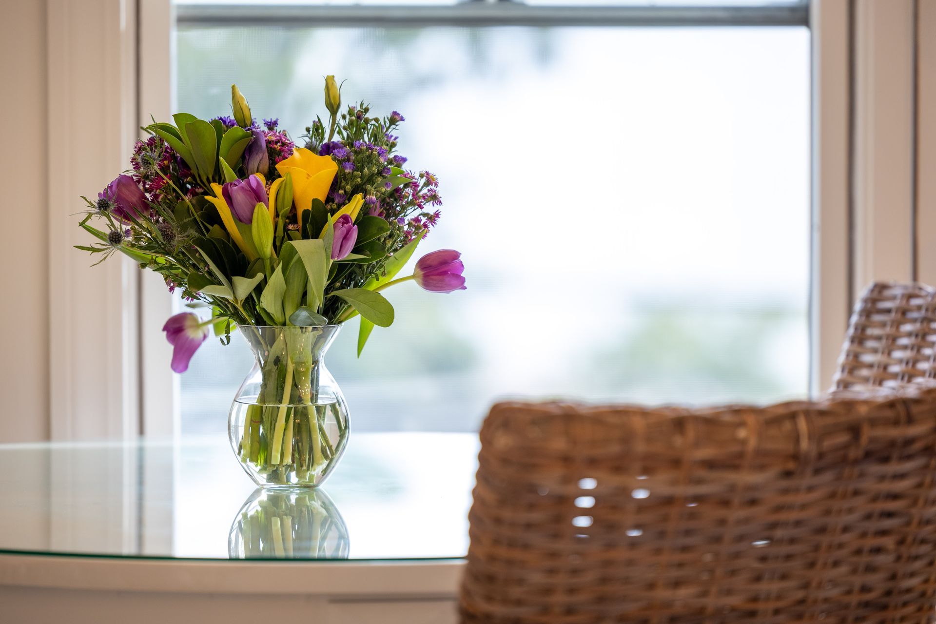 Bouquet of purple, yellow, and green flowers in vase on glass table, next to a wicker chair and a window.