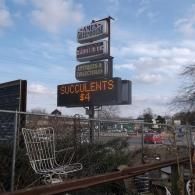 A tall, multi-tiered sign for a greenhouse and antiques shop reads 