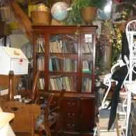 A dark wooden hutch filled with books, surrounded by furniture and decor items in a cluttered room or shop setting.