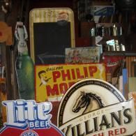 A collection of vintage signs and beer tap handles, including Philip Morris and George Killian's, in a dim, rustic setting.