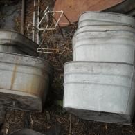 A stack of three galvanized steel tubs sitting outdoors on dry leaves and dirt, with a wire frame nearby.