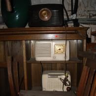 Three vintage radios displayed on wooden shelving, including one black unit on top and two cream-colored units below.