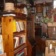 A cluttered antique shop with a wooden bookshelf filled with books, chairs stacked in the background, and wicker baskets.