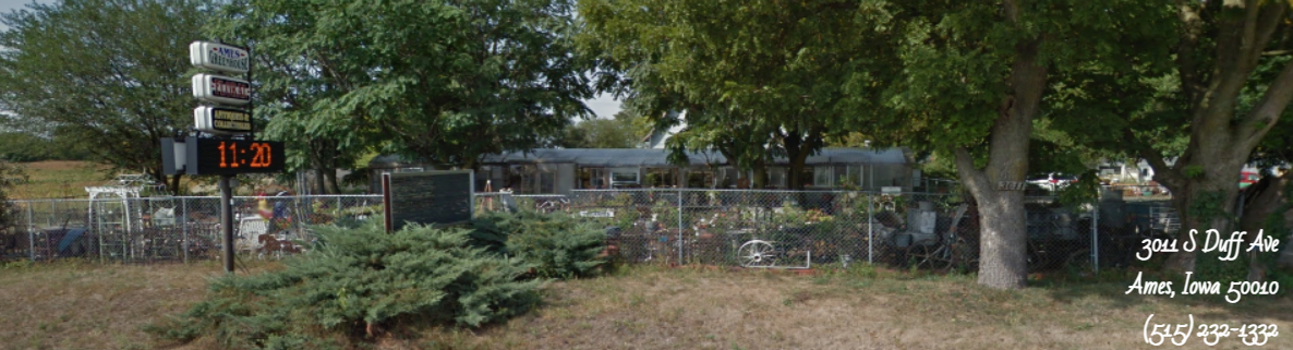 A wide shot of a fenced property with a digital time sign reading 11:28, surrounded by trees and a one-story building.