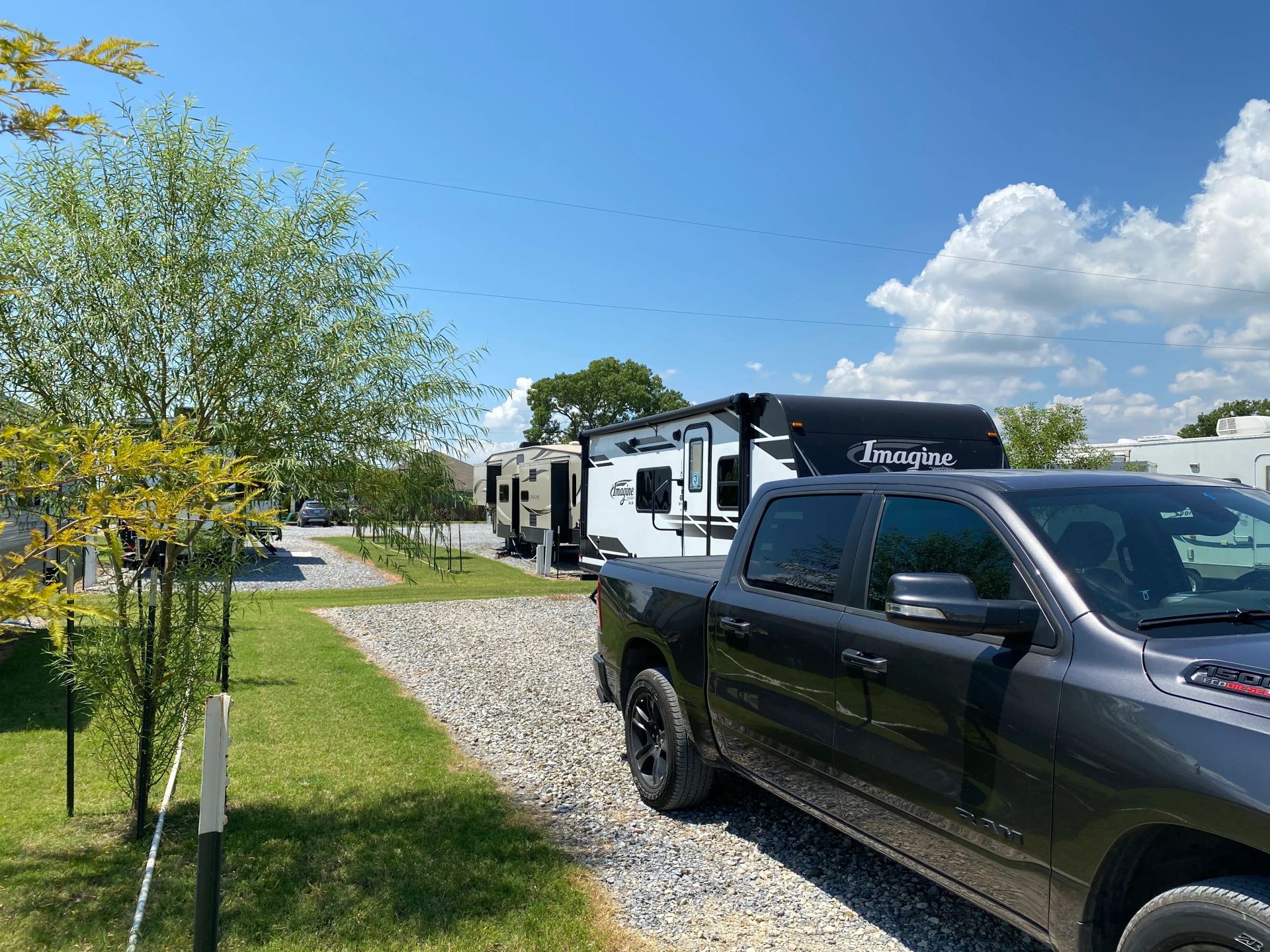 A dark pickup truck parked next to a gravel RV campsite with RVs and trees on a sunny day.