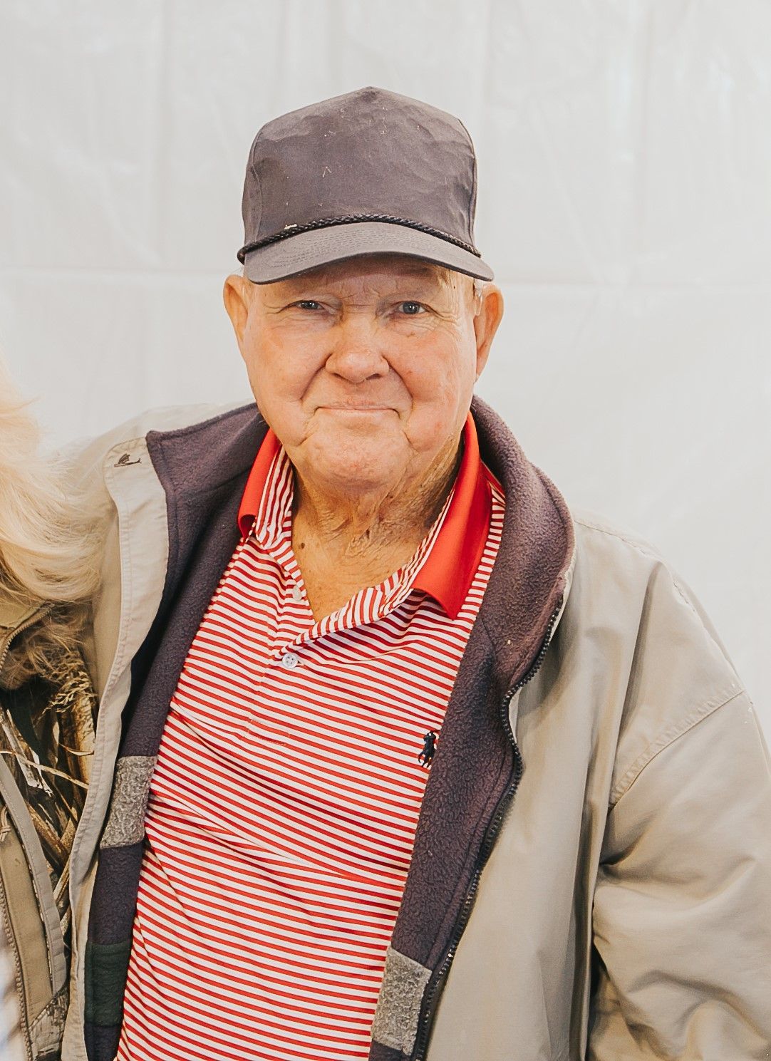 A person wears a dark cap, red and white striped polo shirt, and a light-colored jacket, looking forward against a wall.