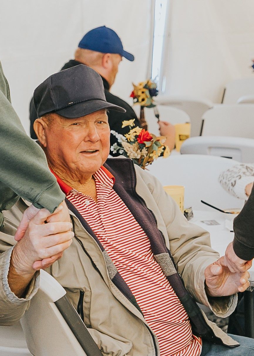 An older person wearing a baseball cap and striped shirt sits at a table, holding hands with others in a bright setting.