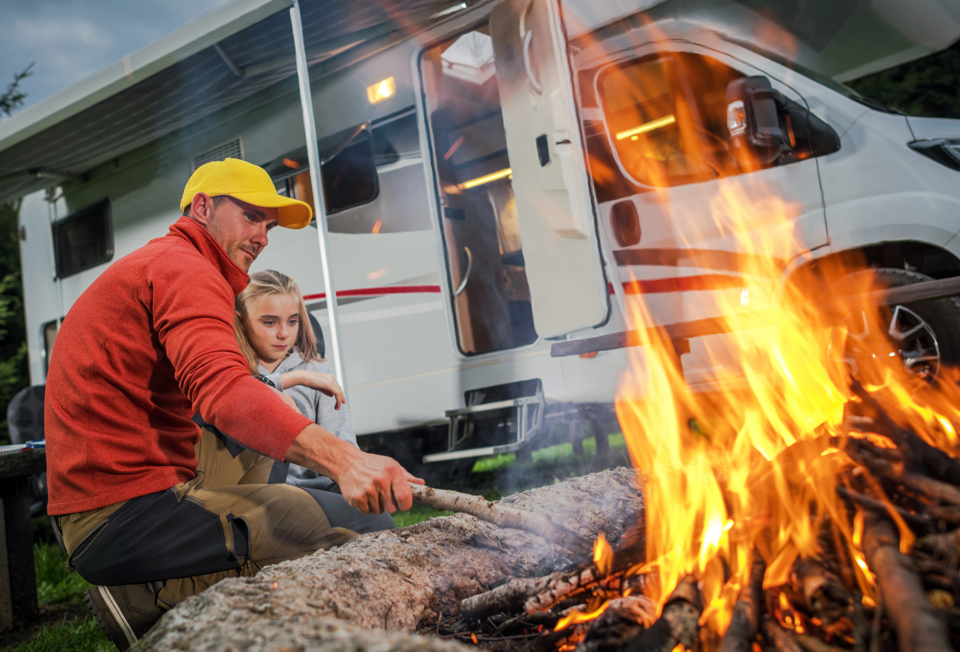 A man and a child are roasting marshmallows over a campfire in front of a rv.