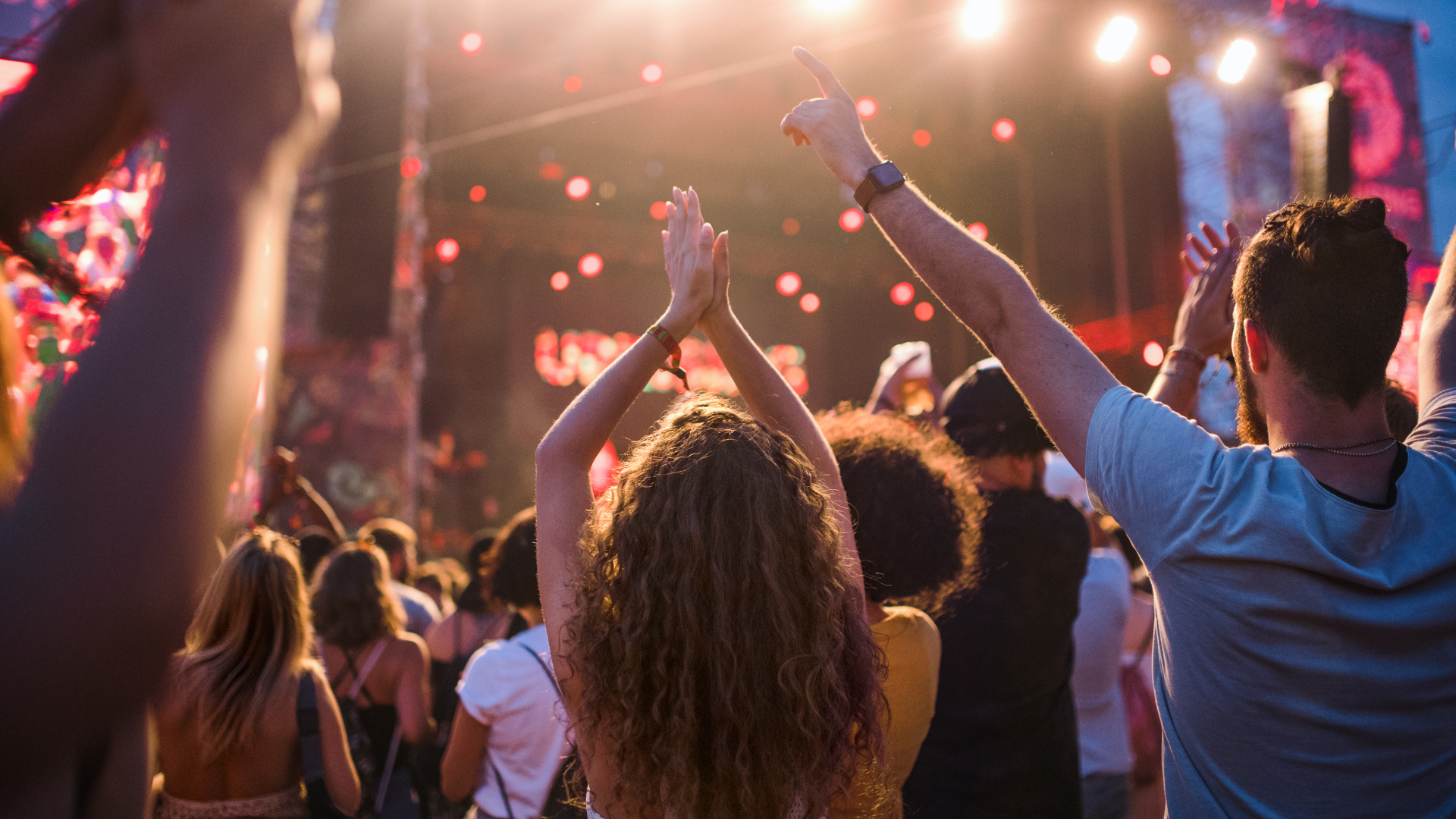 People with arms raised, clapping at an outdoor music concert. Stage lights glow brightly.