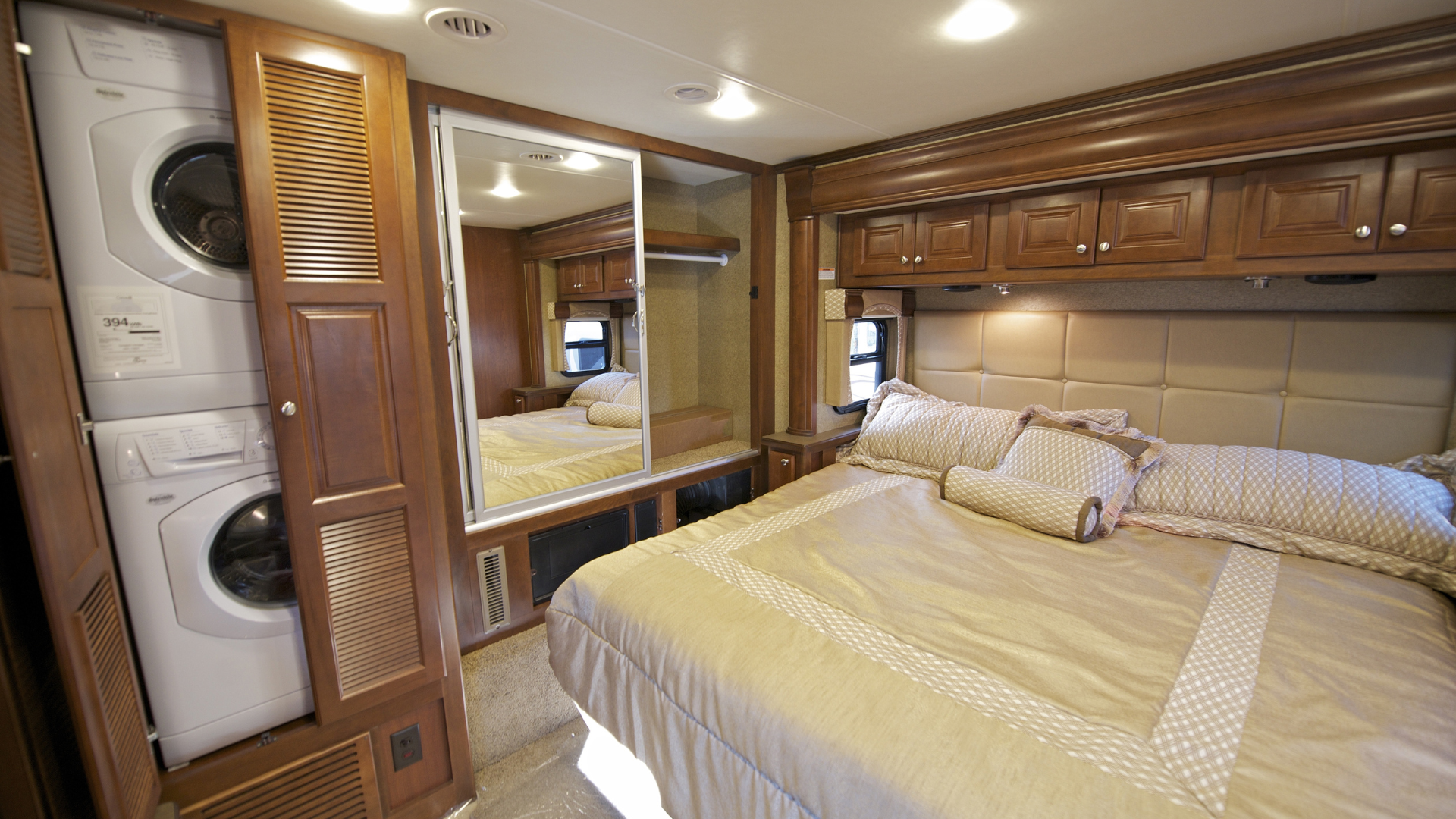 Bedroom in a recreational vehicle with a washing machine, bed, large mirror, and wooden cabinetry.
