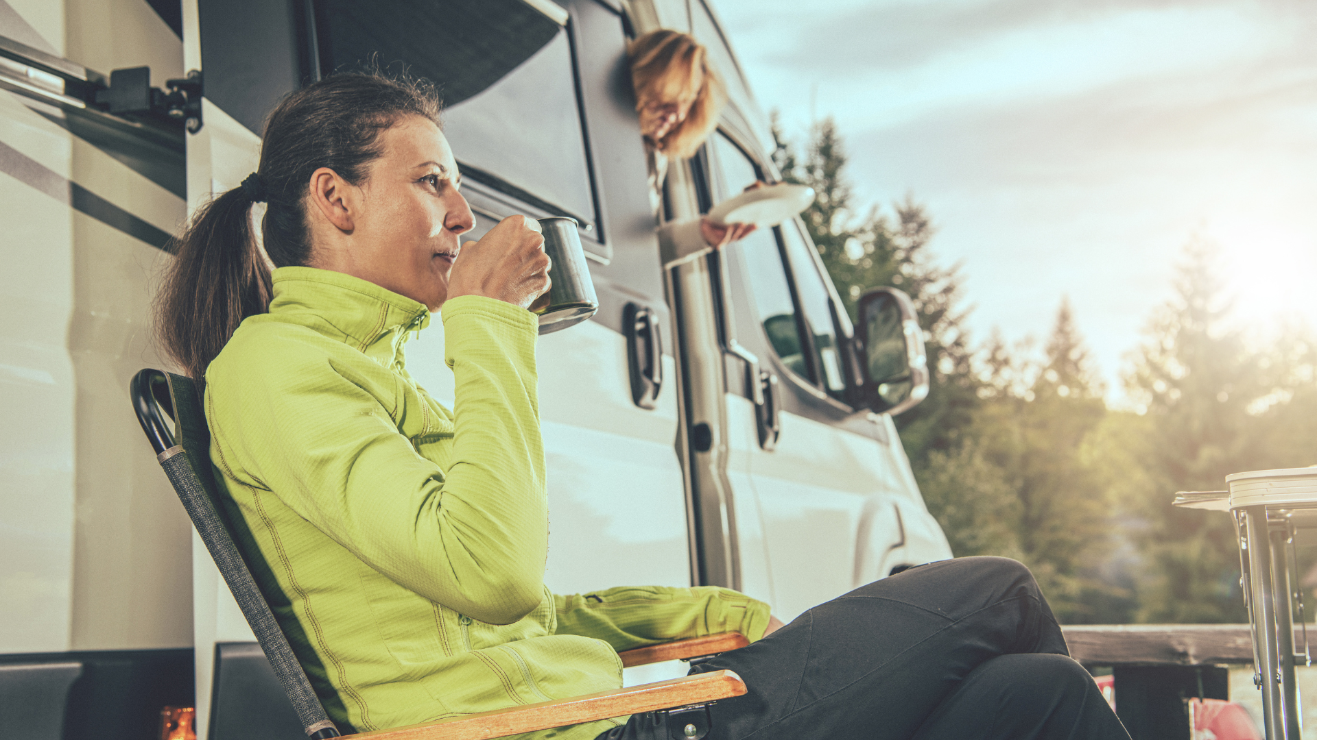 Woman drinking coffee outside RV, another person inside holding a plate, sunny outdoor setting.