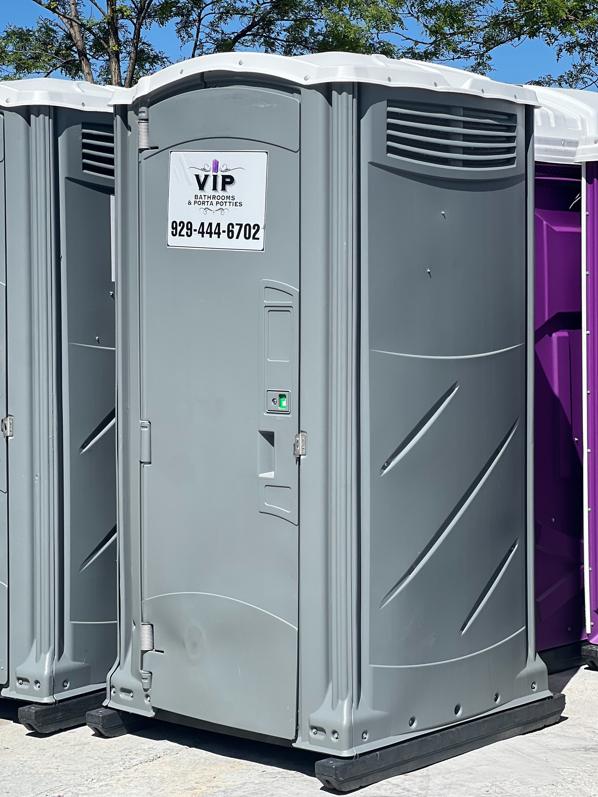 Gray portable toilet with a VIP sign, outdoors beside a purple unit on a sunny day