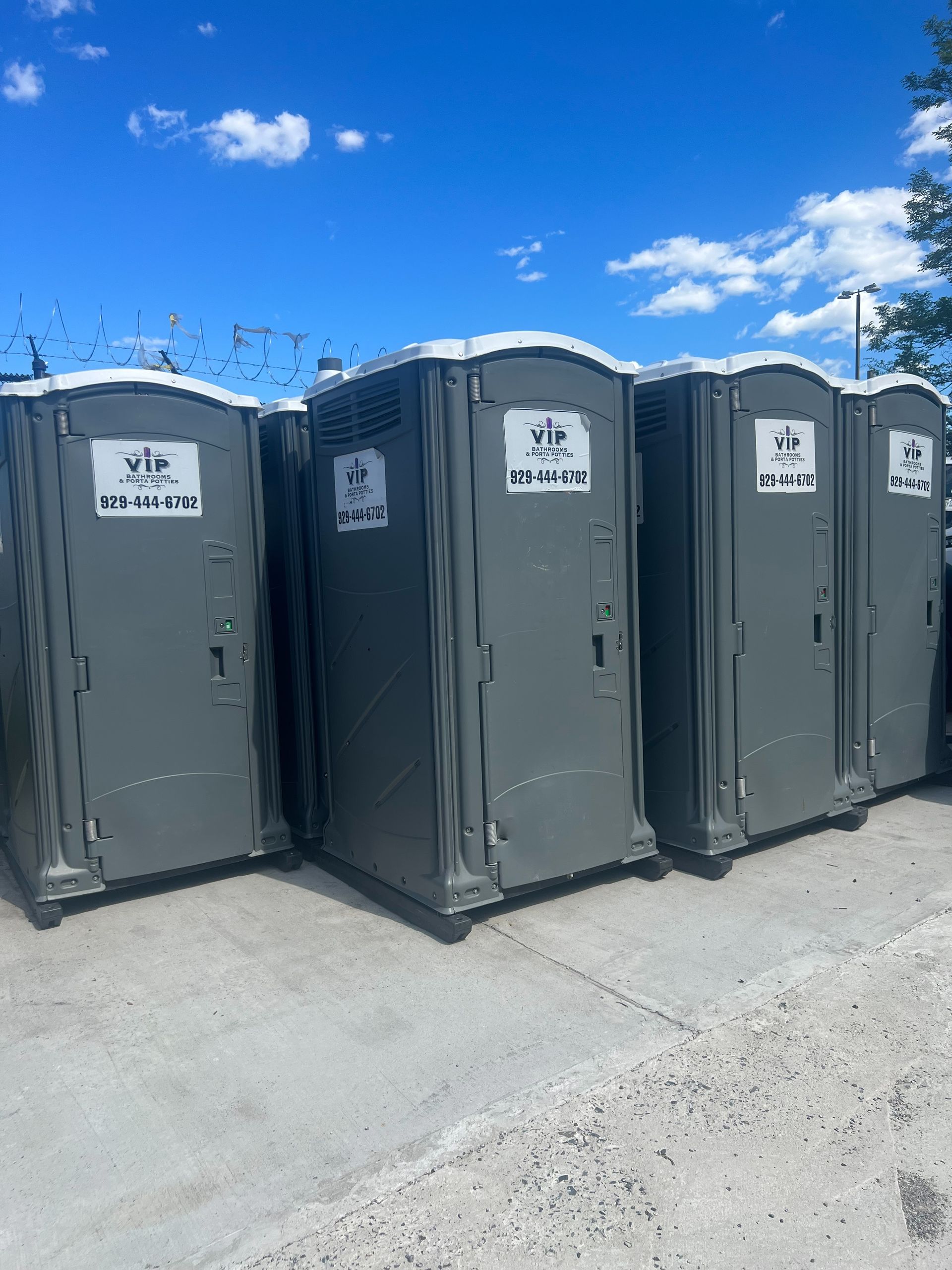 Four gray portable toilets lined up outdoors under a bright blue sky.