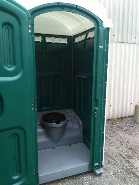 Open portable toilet with green door and gray interior on gravel outside a corrugated building