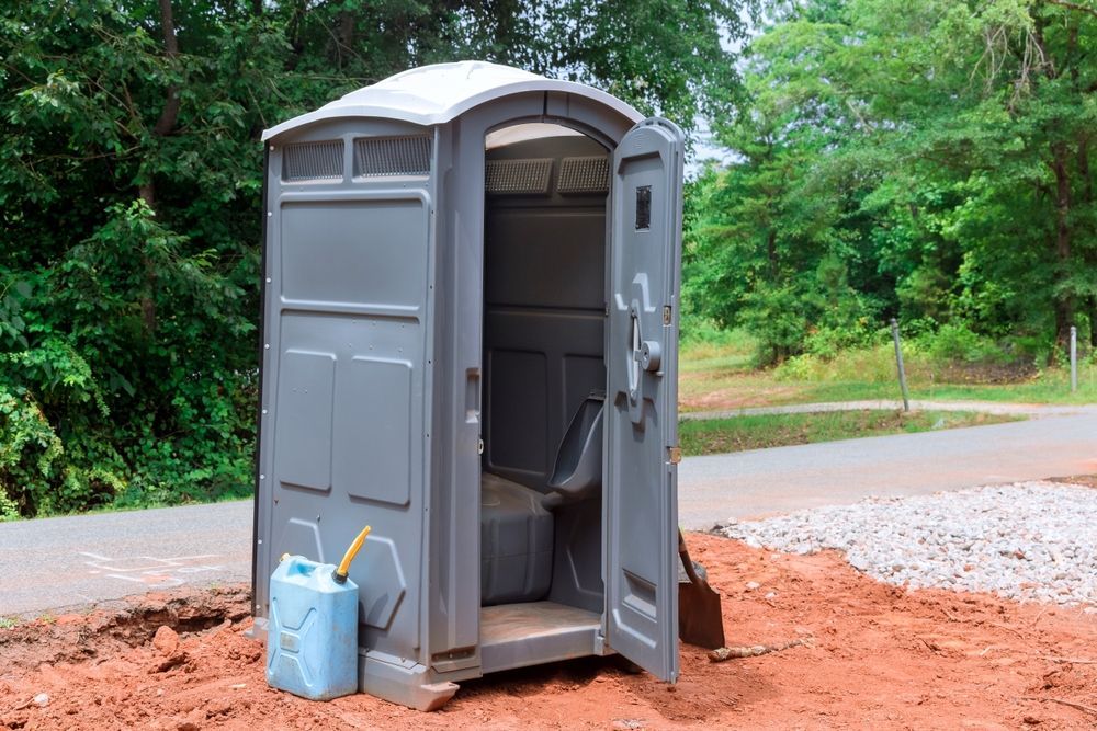Open portable toilet beside a road, with a blue cleaning bucket on red dirt.