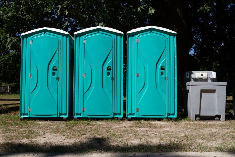 Three teal portable toilets beside a gray outdoor handwashing station on grass.