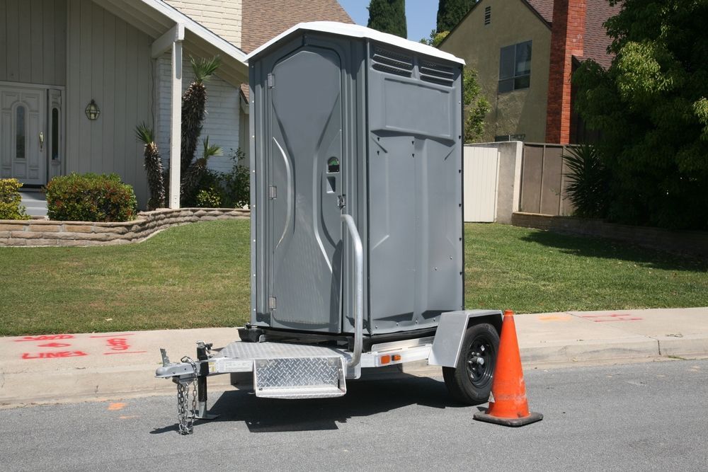 Portable gray toilet on a trailer parked on a suburban street beside an orange traffic cone.