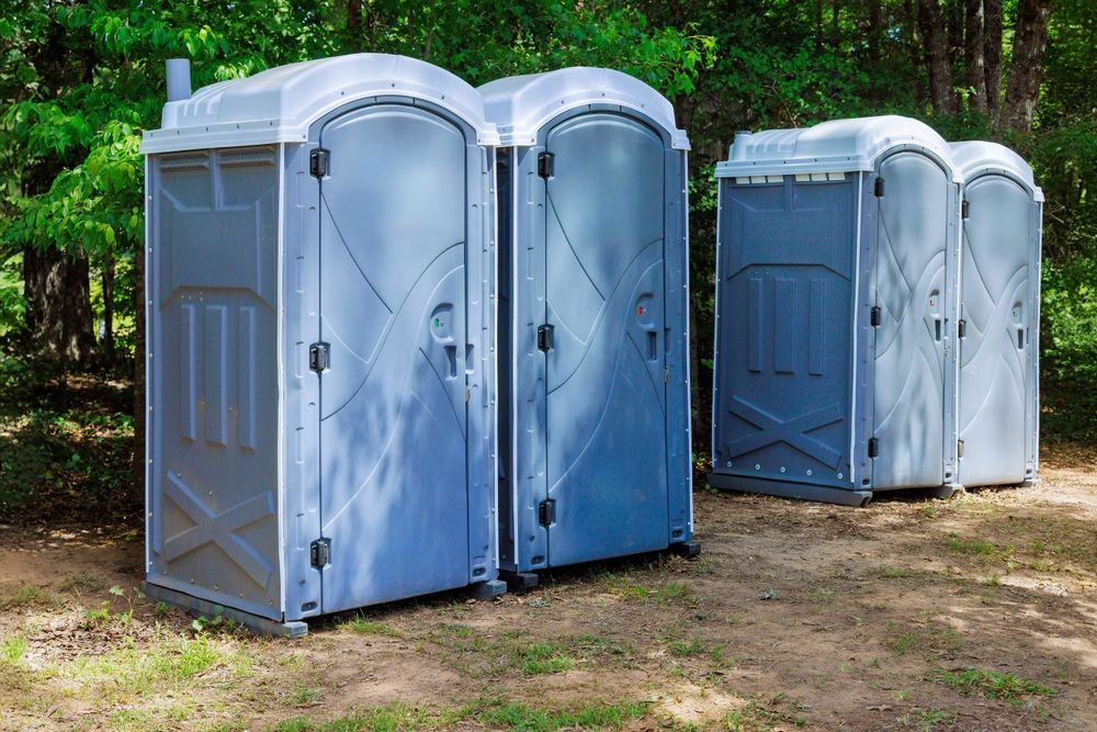 Four blue portable toilets on a dirt path beside trees