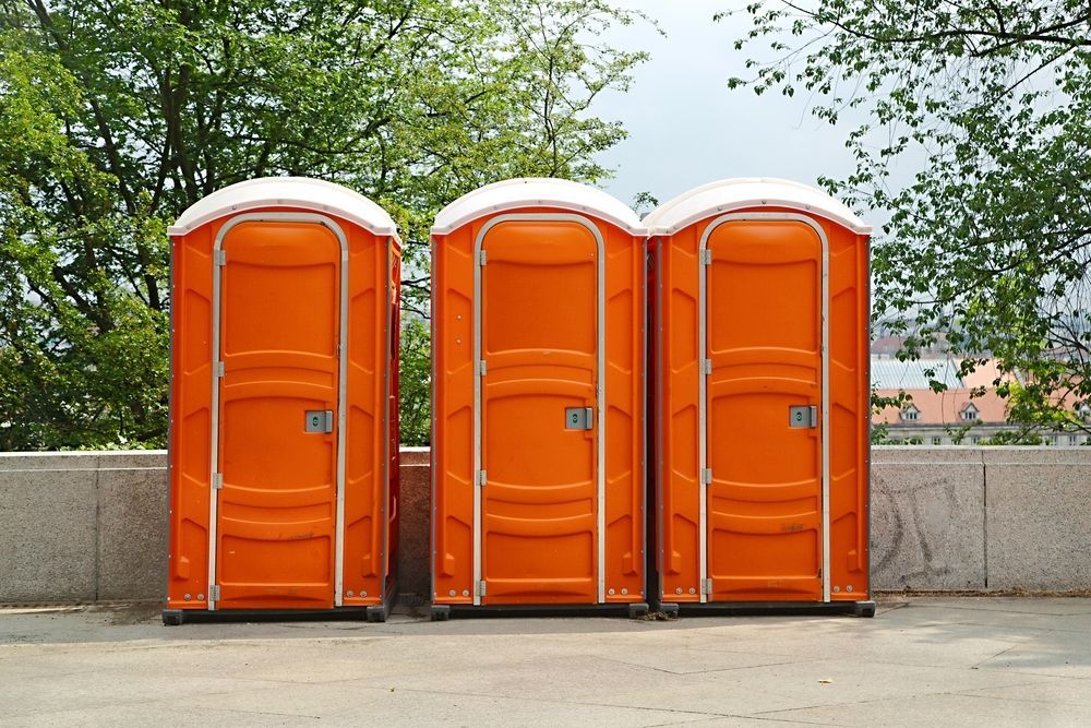 Three orange portable toilets lined up outdoors beside a concrete wall and trees