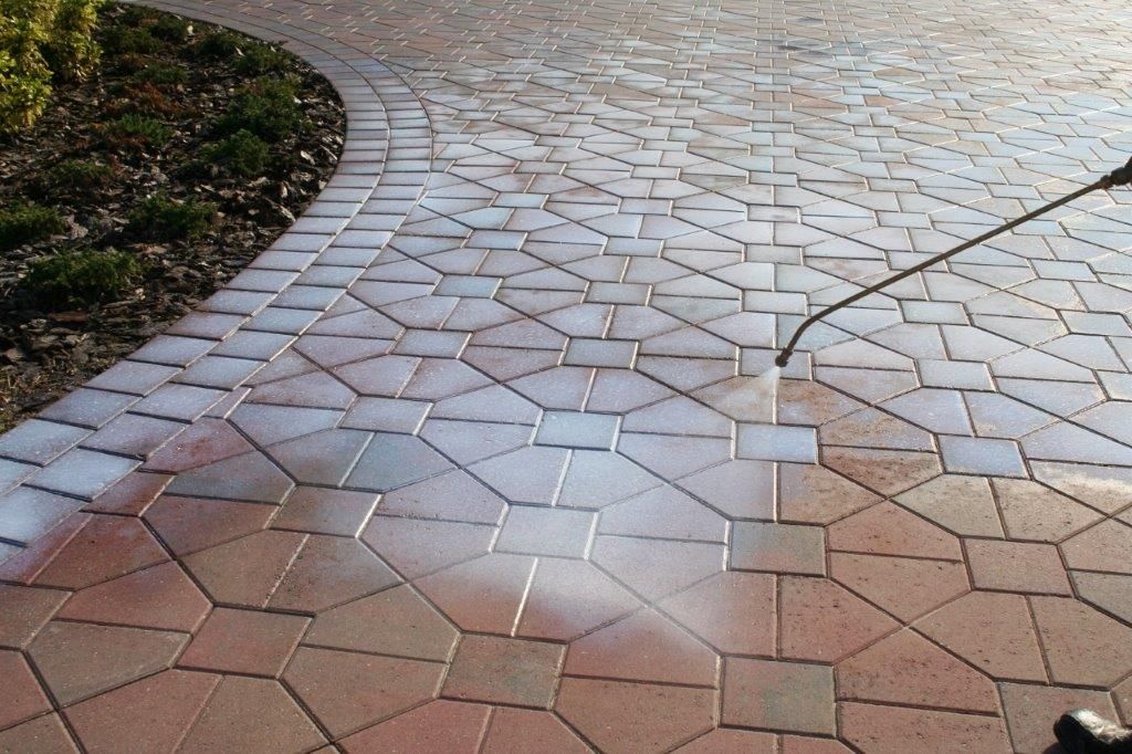 A person sprays sealant onto a patterned brick driveway, showing a contrast between treated and untreated areas.