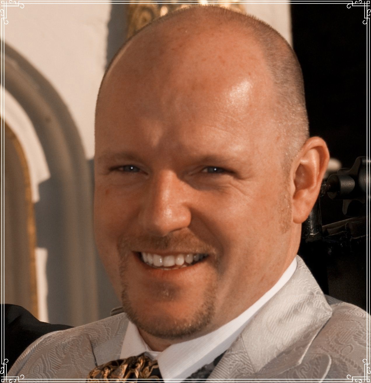 Man with bald head smiles, wearing a patterned jacket, outdoors.