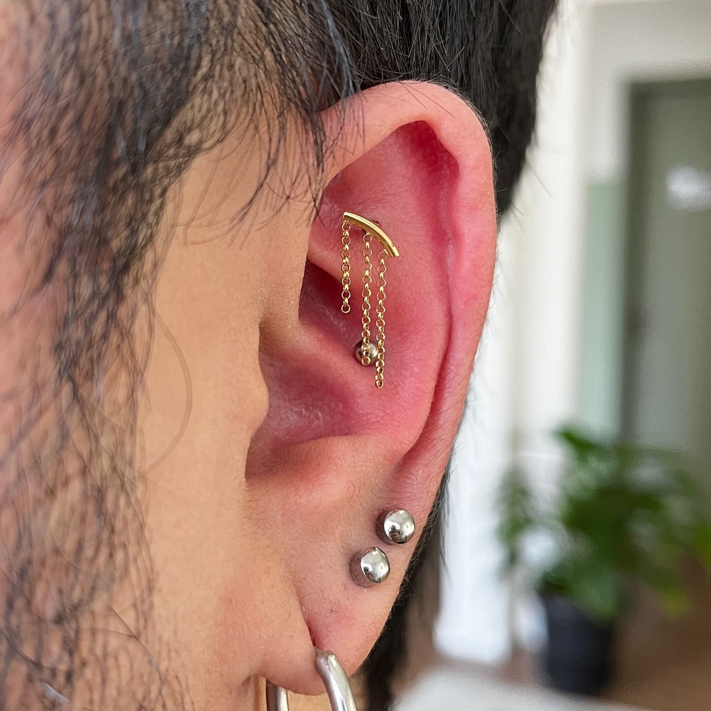 Detalhe da orelha com múltiplos piercings, incluindo um brinco de corrente dourada com contas penduradas.