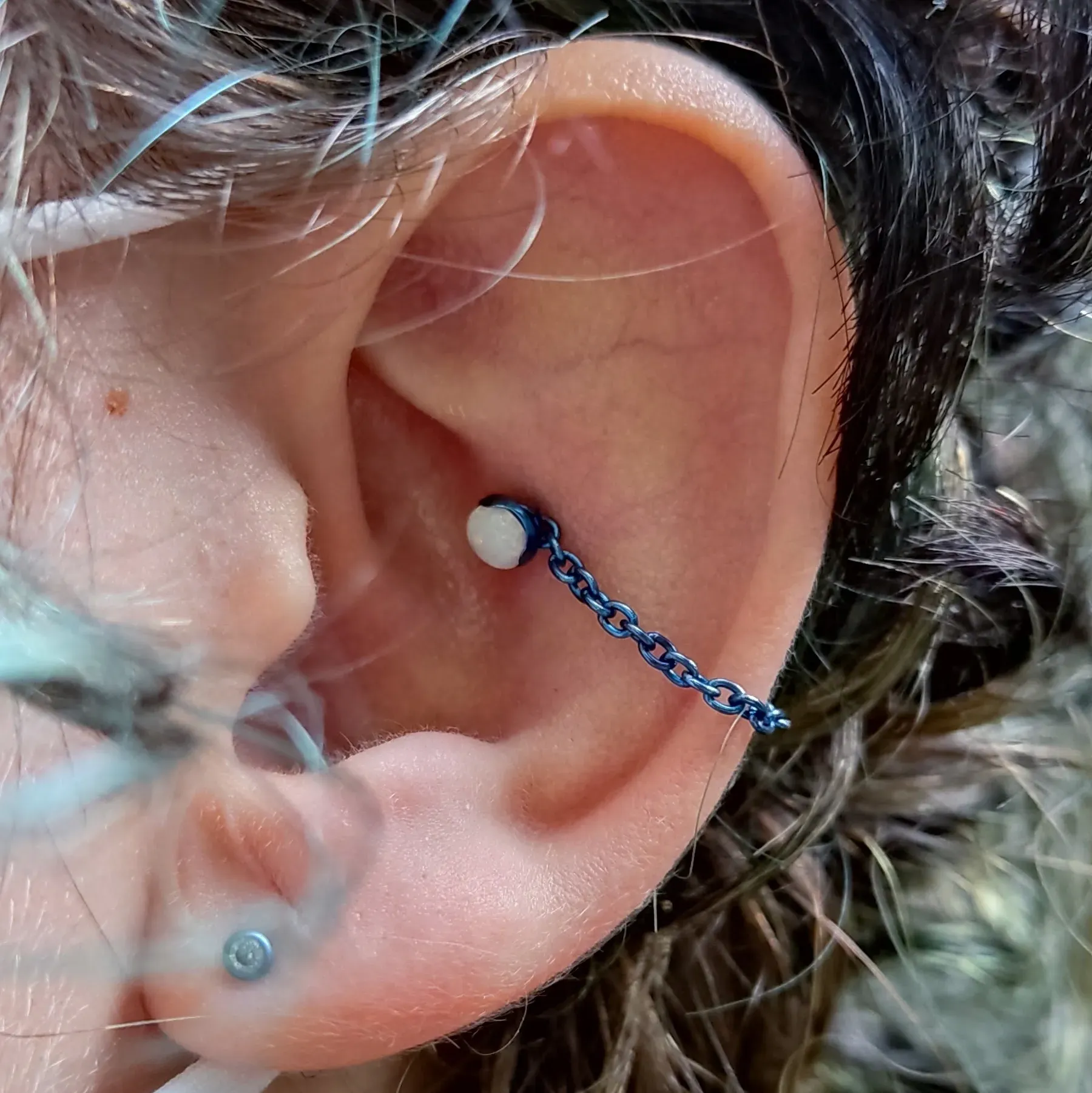 Detalhe da orelha com um brinco de corrente azul conectando dois piercings, um na hélice e outro no lóbulo.