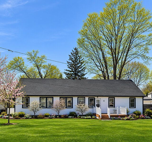 White single-story house with black roof, flower beds, and spring trees under a blue sky