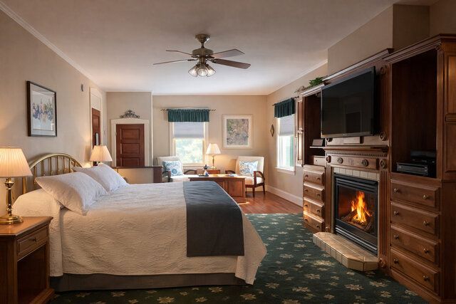 Cozy hotel bedroom with fireplace, king bed, dresser, TV, and a sitting area by a window.