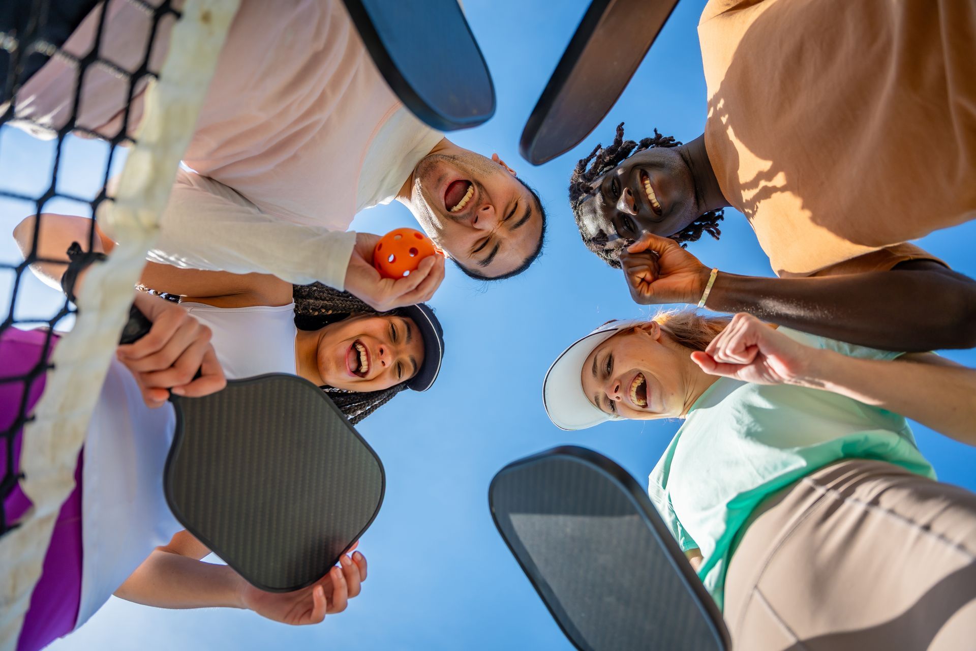 People holding pickleball paddles huddle in a circle under a blue sky, smiling down at the camera.