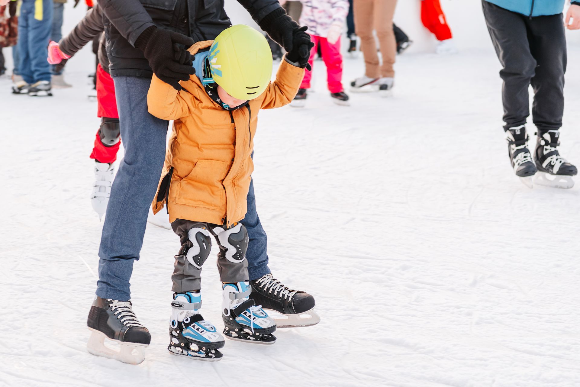Adult helping child ice skate on an outdoor rink, both wearing winter clothes and helmets