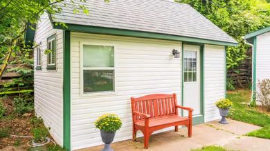 Small white garden shed with gray roof, red bench, and potted plants in a leafy backyard.