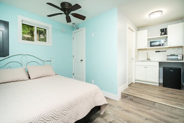 Light blue bedroom with a bed, ceiling fan, window, and small kitchenette