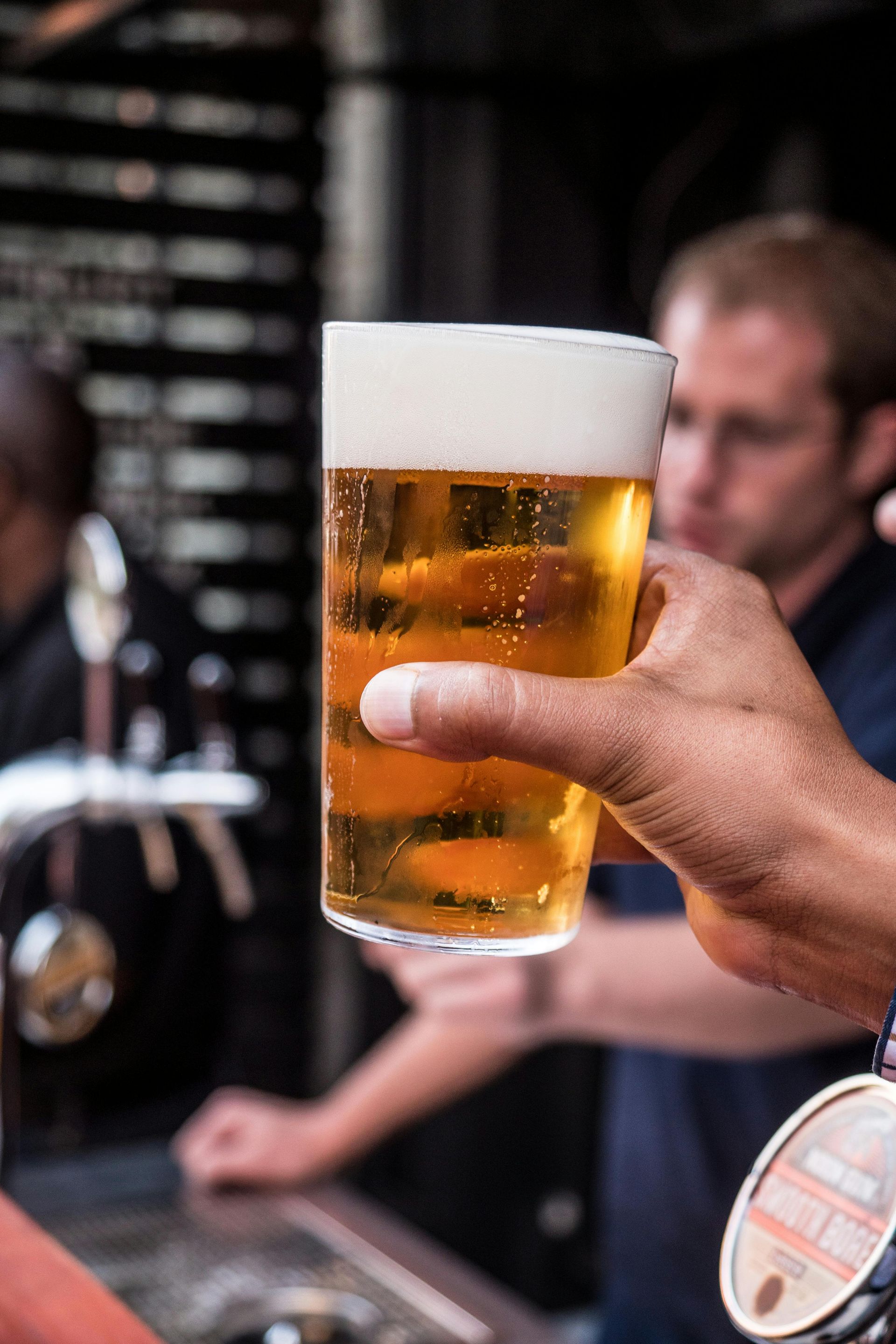 Hand holding a foamy pint of beer at a bar, with blurred people in the background