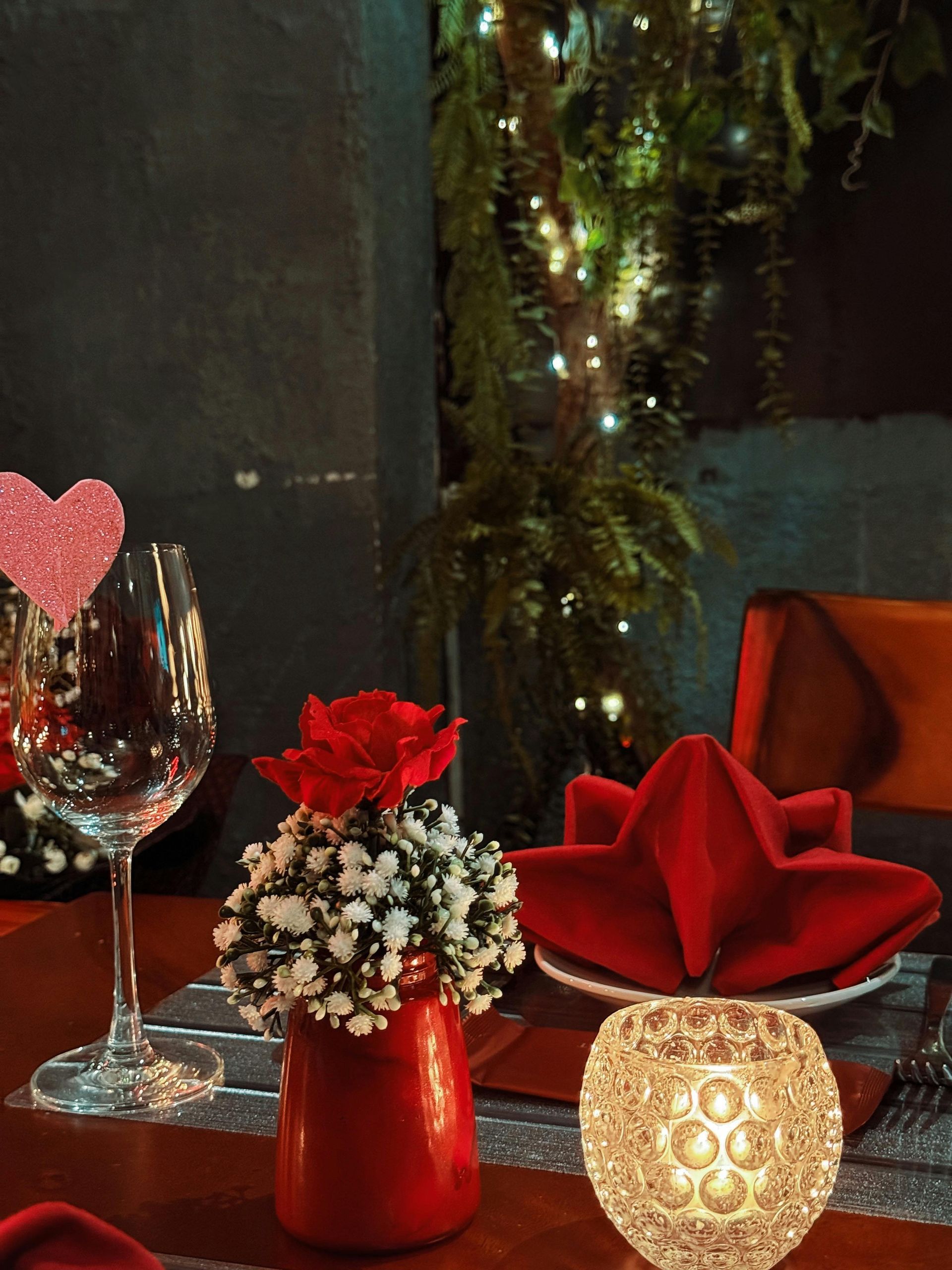 Romantic table setting: red rose centerpiece, wine glass, napkin, candle, fairy lights, and pink heart.