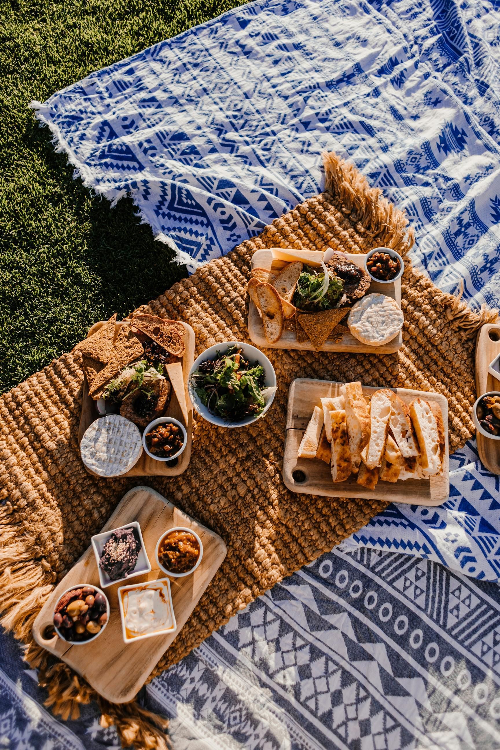Picnic spread on a blanket: wooden boards with food like bread, dips, and a bowl of green relish.