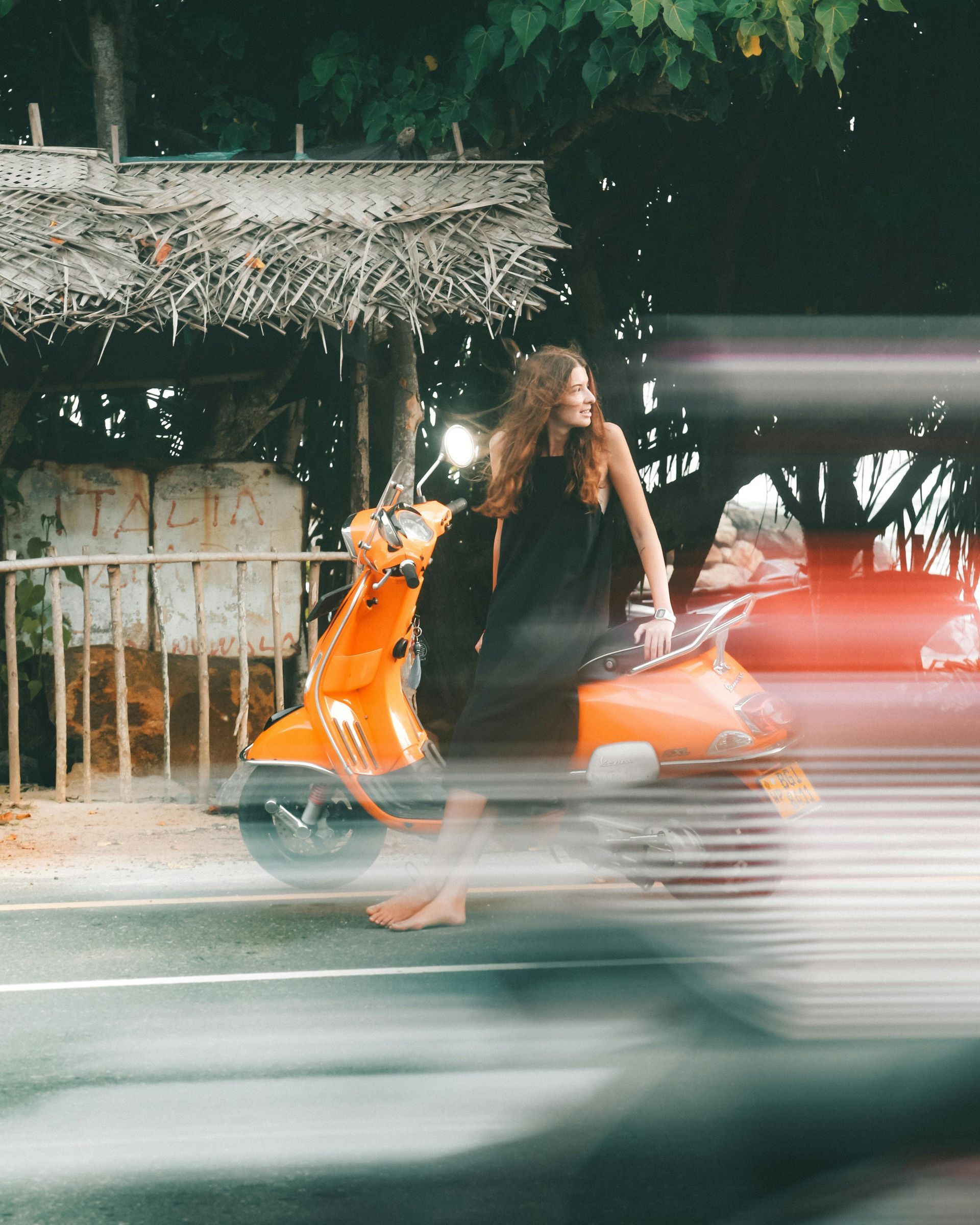 Woman standing beside an orange scooter on a roadside, with motion-blurred traffic passing in front.
