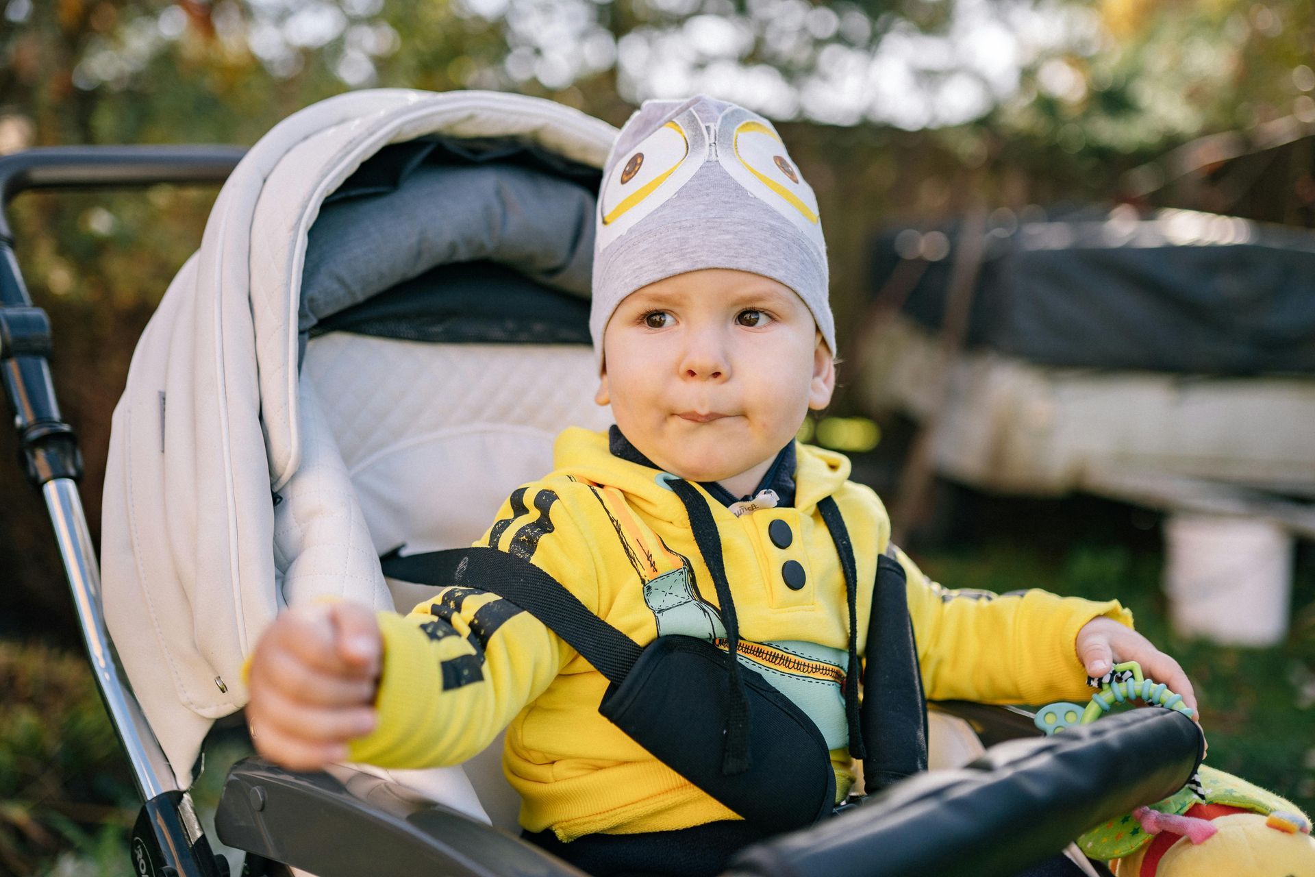 Child in a yellow jacket sitting in a stroller outdoors, holding a hand up.