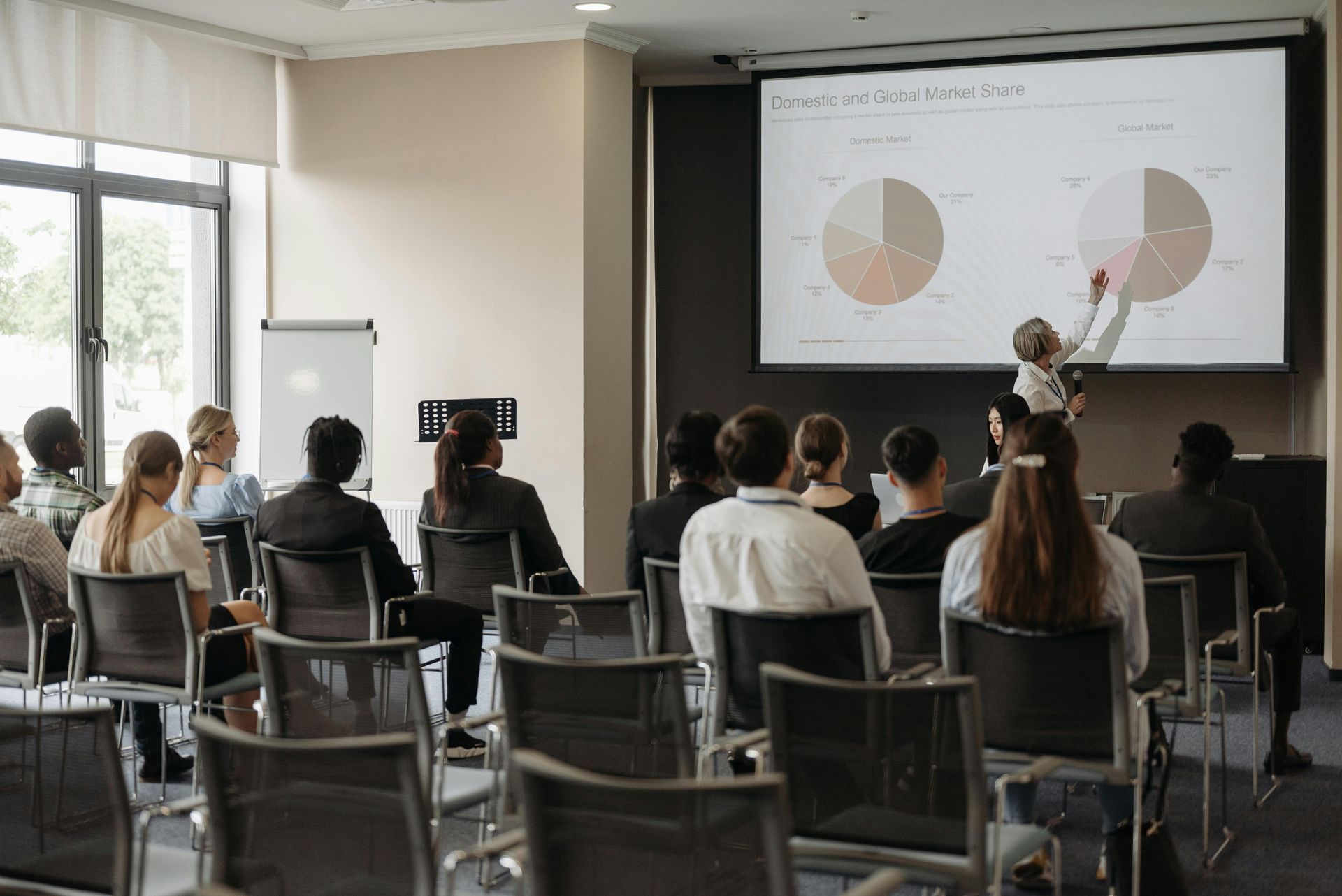 Meeting room with seated audience watching a presenter explain charts on a projected screen.