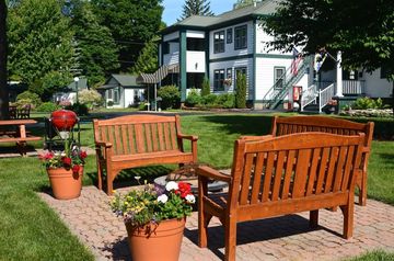 Two wooden benches on a garden patio with potted flowers, in front of a house