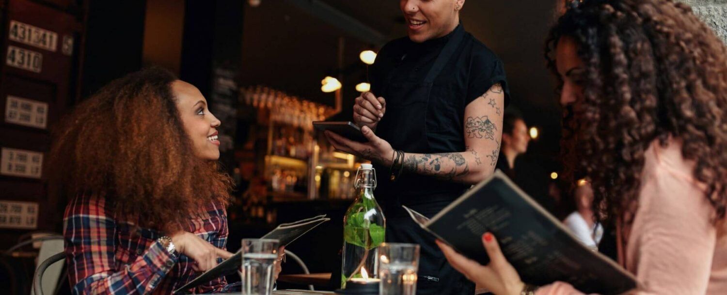 Friends chatting at a café table while a server holds a drink order