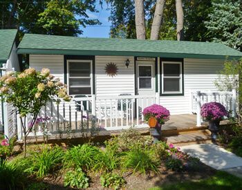 Small white house with green roof, front porch, flowers, and a walkway in a sunny garden.